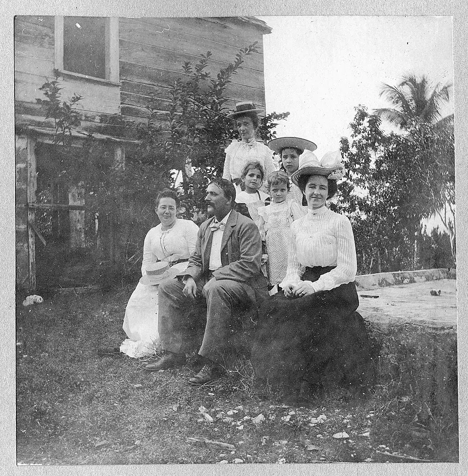 Seven members of a family outside a weathered wooden house with palm trees behind them - a faded, dusty black and white print cleaned up and brought back in color