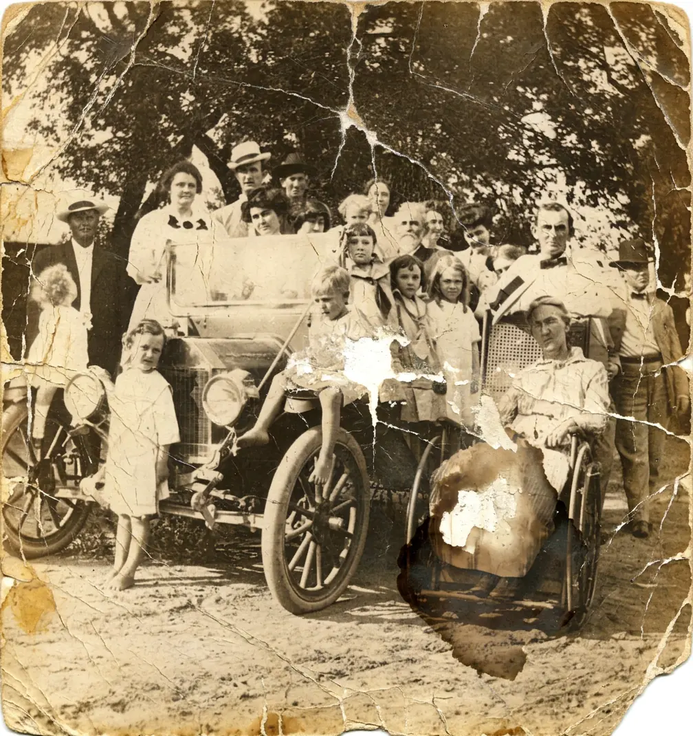 A large family gathered around an early motor car, one of them seated in a wheelchair - deep cracks, torn edges and two holes through the print repaired, and the whole scene back in color